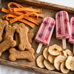Natural homemade dog treats — bone biscuits, sweet potato chews, frozen berry pops and apple chips on a wooden tray.